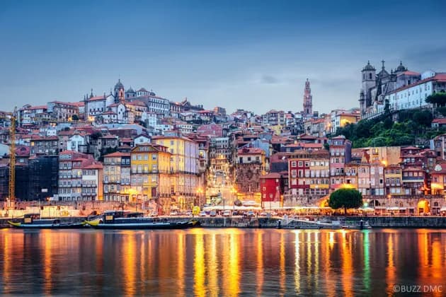 Un denso paisaje urbano de edificios coloridos en una colina, iluminado al atardecer con reflejos en el río en primer plano.