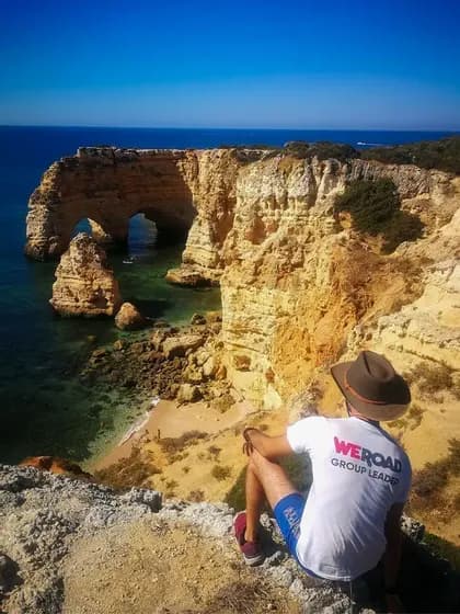 Un capo gruppo WeRoad con un cappello siede su una scogliera che si affaccia su una costa rocciosa con archi naturali sul mare e una piccola spiaggia.