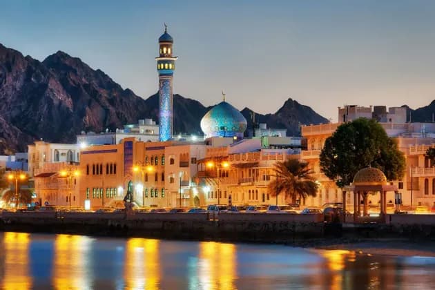 Una moschea illuminata e uno skyline cittadino su un lungomare al tramonto, con aspre montagne che si ergono dietro gli edifici.