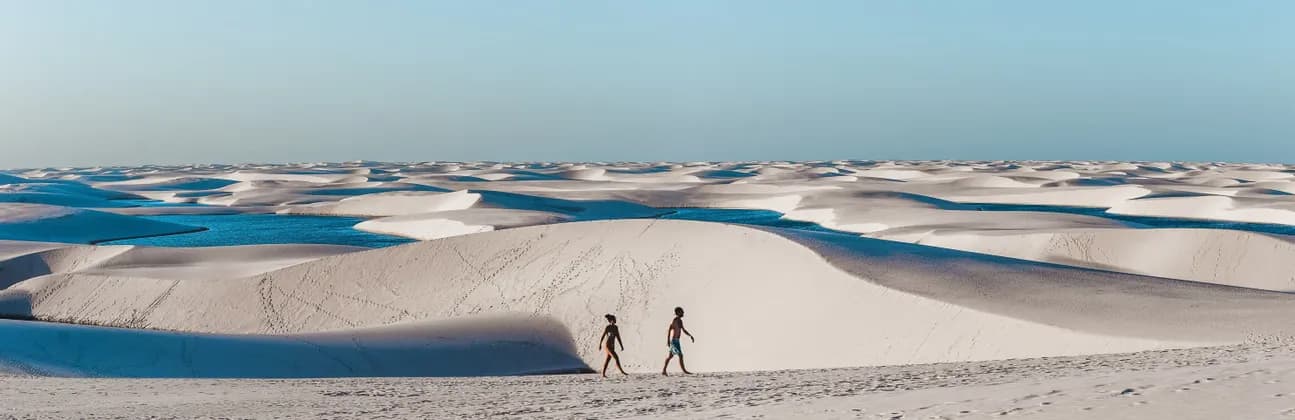 Two people walk across vast white sand dunes dotted with blue lagoons under a clear sky.