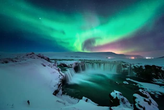 L'aurora boreale verde brilla in un cielo notturno stellato sopra un'ampia cascata in un paesaggio innevato.