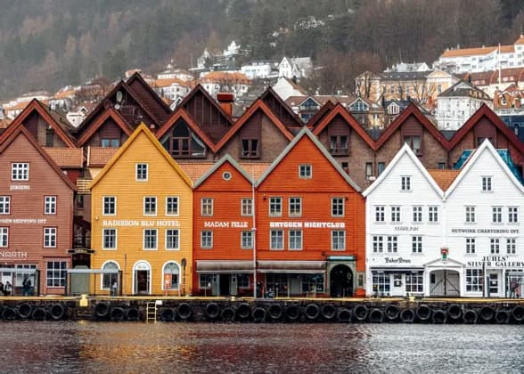 Colorful historic wooden buildings with gabled roofs line a waterfront pier against a hillside background.