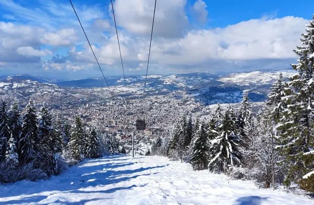 Una funivia scende da una montagna innevata, con vista su una città estesa nella valle sottostante, sotto un cielo parzialmente nuvoloso.