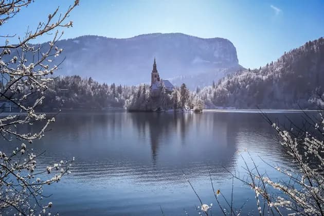 A snow-covered church on an island reflects on a calm lake, with forested mountains in the background under a clear blue sky.