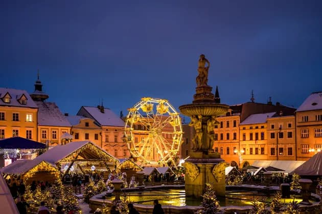 Una noria iluminada y puestos de mercado festivos rodean una fuente de piedra iluminada en la plaza de un pueblo cubierto de nieve al atardecer.