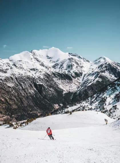 Un skieur en veste orange glisse sur une pente enneigée face à d'immenses montagnes enneigées sous un ciel bleu dégagé
