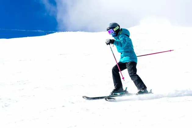 Un esquiador con chaqueta turquesa, casco y gafas esquía cuesta abajo en una brillante pendiente nevada bajo un cielo azul.