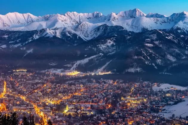 An illuminated town glowing at dusk beneath snow-covered mountain peaks under a clear blue sky.