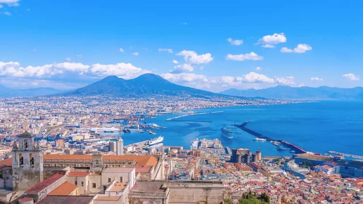 Elevated panoramic view of a coastal city harbor and bay with a large twin-peaked mountain under a blue sky.