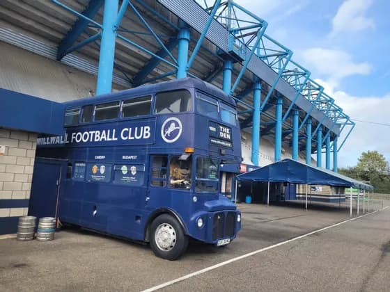 Un autobus a due piani vintage blu di una squadra di calcio parcheggiato fuori da uno stadio con travi del tetto in acciaio blu.