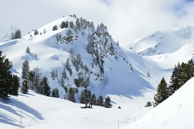 Sciatori e snowboarder che riposano su un'ampia pista innevata circondata da pini e cime alpine