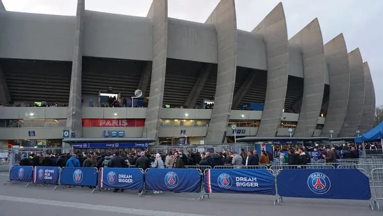 Una folla di persone si riunisce fuori dall'ingresso di un grande stadio in cemento delimitato da barriere blu.