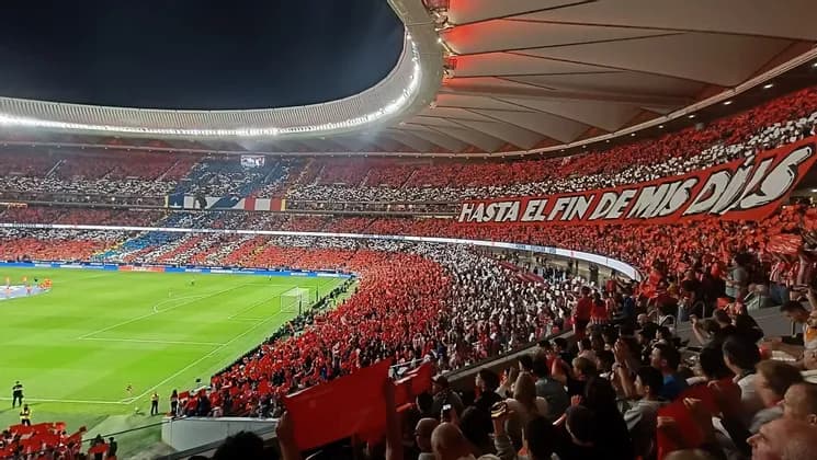 Uno stadio affollato pieno di tifosi festanti che tengono cartelli rossi attorno a un campo da calcio verde illuminato da luci vivaci