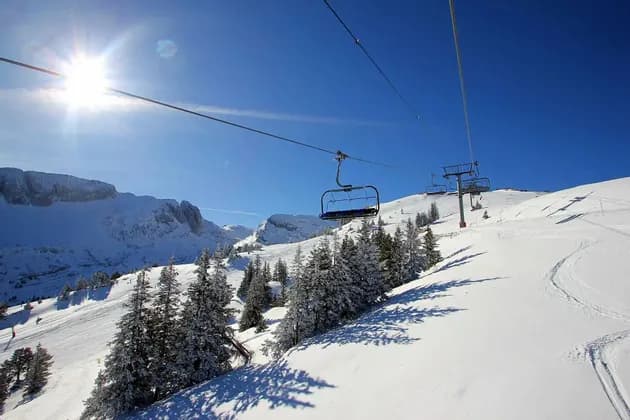 Un telesilla vacío asciende por una ladera de montaña cubierta de nieve, rodeada de pinos en un día soleado.