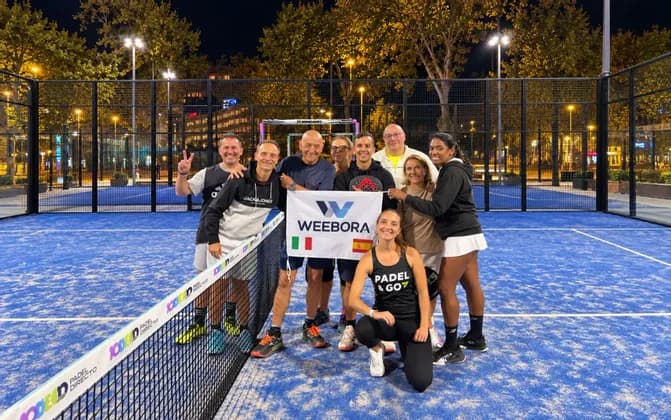 Un gruppo di sorridenti giocatori di padel in posa con uno striscione su un campo da padel blu all'aperto di notte.
