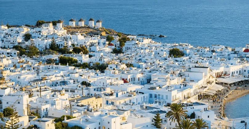 Edifici di una cittadina costiera imbiancata a calce si estendono su una collina che conduce a mulini a vento accanto a un mare blu.
