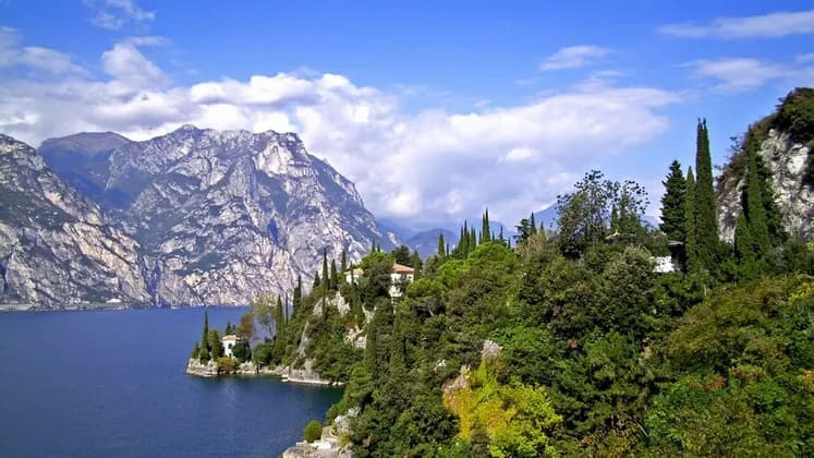 Lush green hills with cypress trees and small houses along a blue lake with steep rocky mountains in the background.