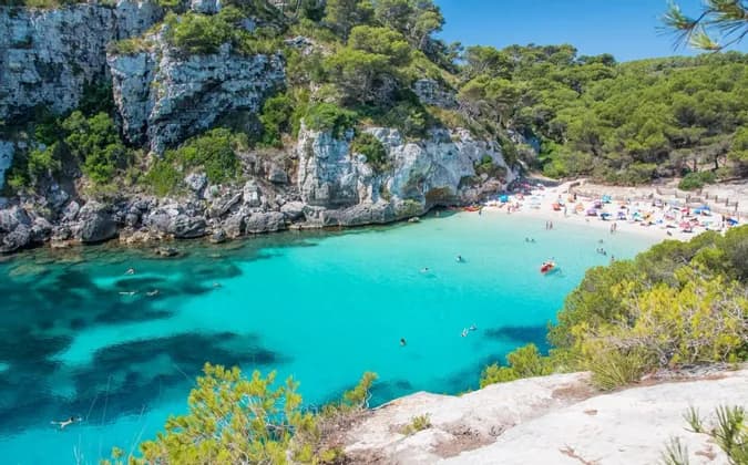 Vue aérienne d'une crique où des personnes nagent dans une eau turquoise claire et se détendent sur une plage de sable blanc entourée de falaises rocheuses et d'arbres.