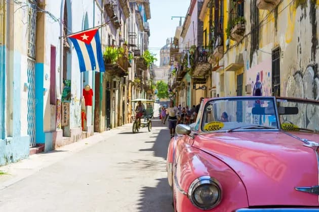 A pink vintage car parked on a narrow, sunlit street lined with colorful buildings, with a Cuban flag hanging from a balcony.