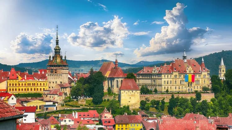 Eine historische Stadt mit einem zentralen Glockenturm und farbenfrohen Gebäuden mit roten Dächern, vor dem Hintergrund grüner Hügel und blauem Himmel.