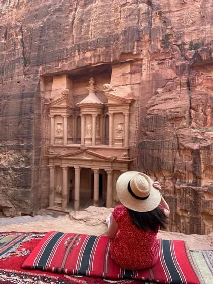 Une femme en robe rouge et chapeau de paille est assise sur un tapis, admirant un grand temple sculpté dans une falaise de roche rouge.