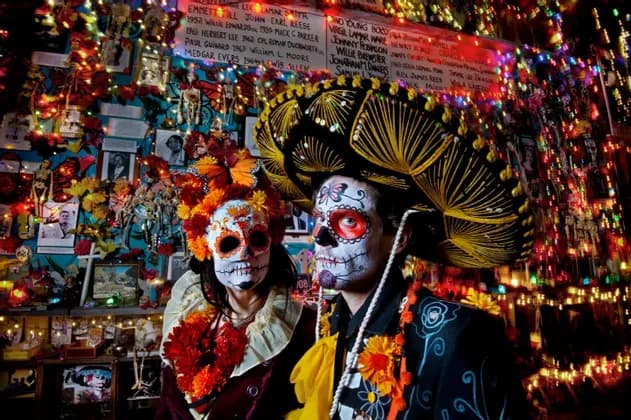Dos personas con disfraces del Día de Muertos y maquillaje de calavera de azúcar posan frente a una pared decorada con luces y fotos.