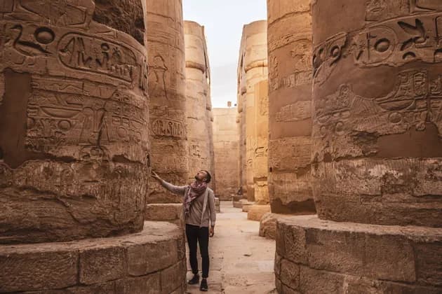 A man stands in an ancient temple hall, looking up at the hieroglyphs carved into the massive stone columns.