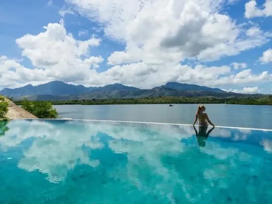 Una donna al bordo di una piscina a sfioro guarda una baia tranquilla verso montagne distanti sotto un cielo parzialmente nuvoloso.