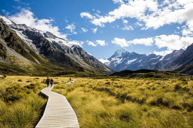 Un voyage de groupe WeRoad se promène sur une passerelle en bois, traversant une vallée verdoyante, avec des montagnes enneigées en fond.