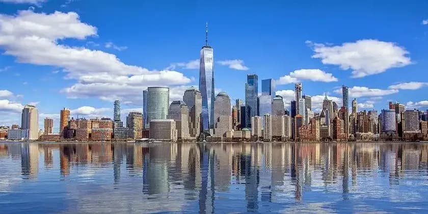 A modern city skyline with tall skyscrapers reflecting in the calm water under a blue sky with scattered clouds.