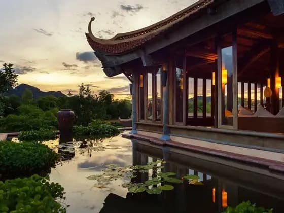 A wooden building with a traditional curved roof overlooks a tranquil pond with water lilies, with lush mountains in the background at sunset.