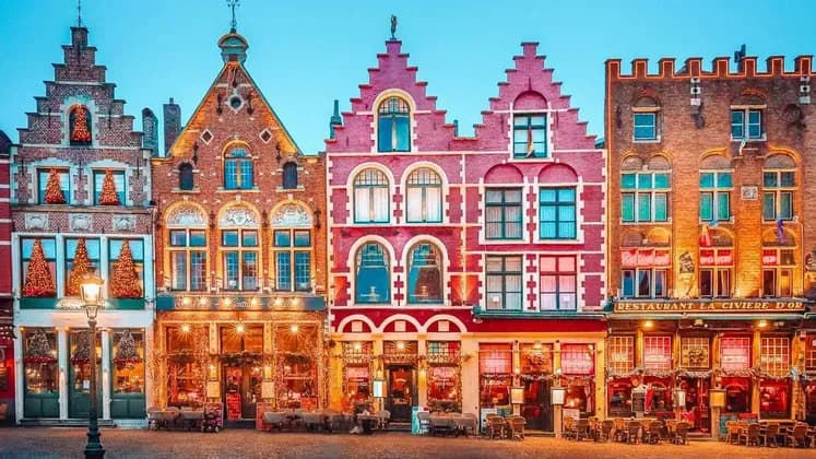 Una hilera de casas de colores, tradicionales y con tejados a dos aguas, decoradas con luces y árboles de Navidad en una plaza adoquinada al anochecer.