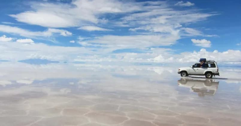 Ein weißer SUV einer WeRoad-Gruppenreise fährt über eine weite, nasse Salzpfanne, die den bewölkten blauen Himmel wie einen Spiegel reflektiert.