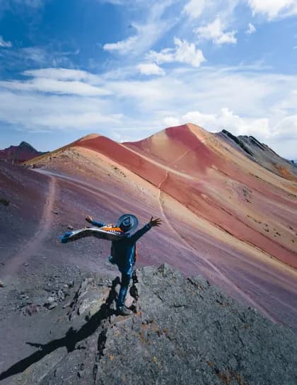 Una persona con sombrero se alza en un promontorio rocoso con los brazos abiertos, contemplando una cordillera de montañas coloridas y a rayas bajo un cielo azul.
