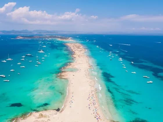 Una vista aerea di una lunga lingua di sabbia che si estende in un mare turchese, con molte barche ancorate e persone sulla spiaggia.
