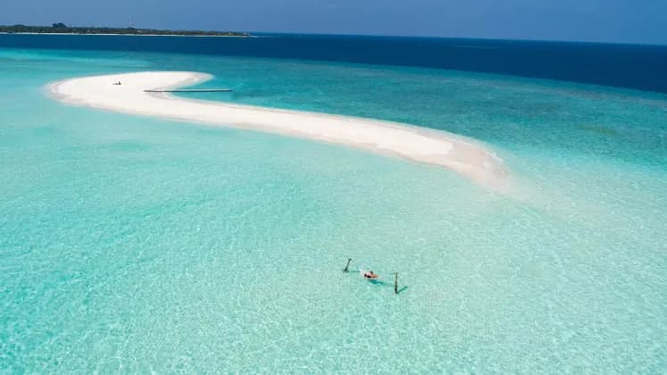 Vista aerea di una persona che galleggia su una zattera in acque cristalline turchesi, accanto a un sinuoso banco di sabbia bianco e all'oceano blu profondo.