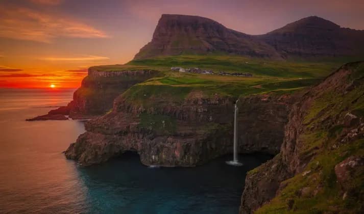 Una cascata si getta da una scogliera erbosa nell'oceano al tramonto, con un piccolo villaggio sulla cima della scogliera.