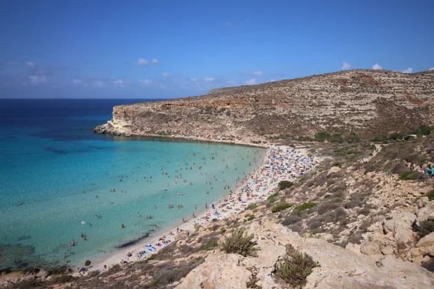 Una vista dall'alto di una spiaggia affollata in una cala rocciosa, con persone che nuotano in acque limpide color turchese sotto un cielo blu.