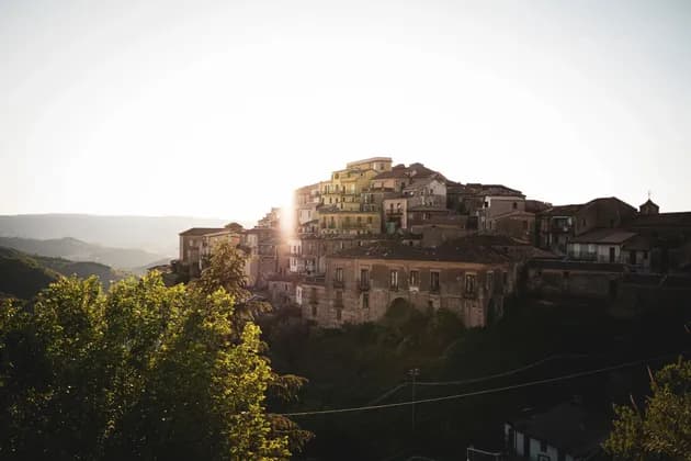 Un borgo di vecchie case costruite su un fianco di collina verdeggiante, con il sole nascente che crea un bagliore luminoso da dietro gli edifici.