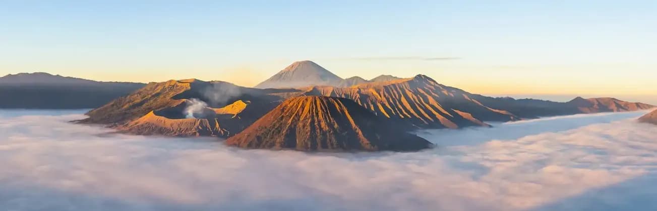 Un volcan fumant et d'autres sommets montagneux s'élèvent au-dessus d'une dense couche de nuages, illuminés par la lumière dorée du lever du soleil.
