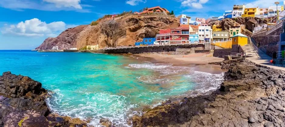 Ein Panoramablick auf ein Küstendorf mit bunten Gebäuden an einer Klippe neben einem Strand mit türkisfarbenem Wasser.