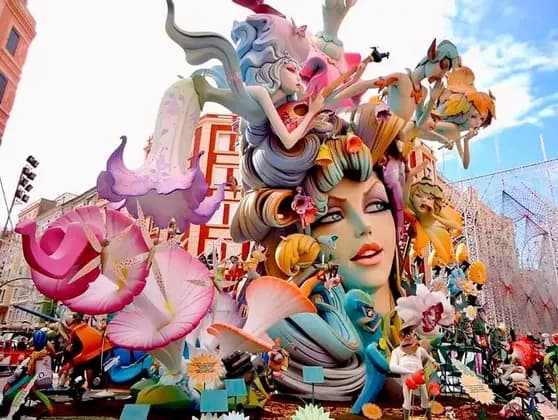 Una scultura imponente e colorata, raffigurante un grande volto femminile, figure fantastiche e fiori, si erge all'aperto in una città.