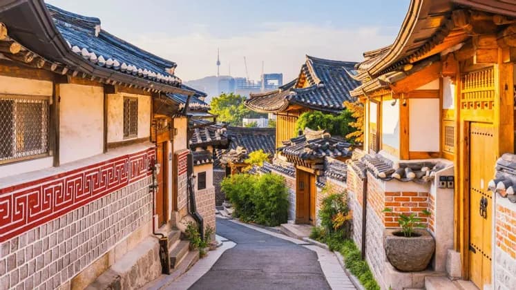 Una stretta via si snoda tra case tradizionali con tetti in tegole, con lo skyline moderno di una città e una torre visibili sullo sfondo.