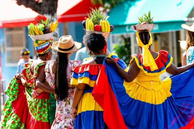 Quattro donne viste di spalle, con indosso abiti tradizionali vivaci e bilanciando cesti di frutta sulla testa mentre camminano.