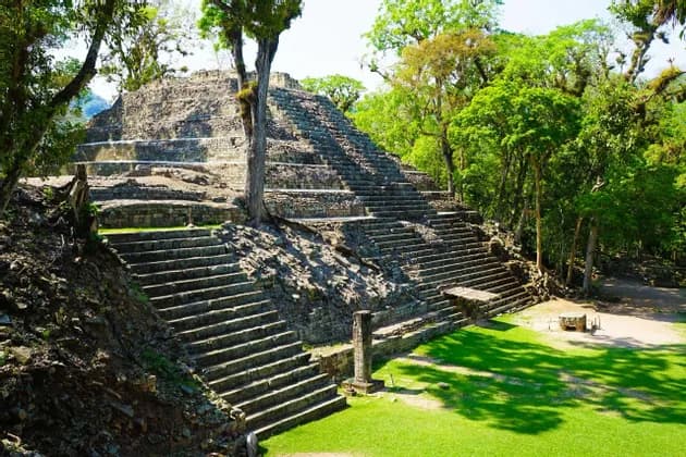 Eine antike Steinstufenpyramide mit steilen Treppen, umgeben von grünen Bäumen und Gras an einem sonnigen Tag.