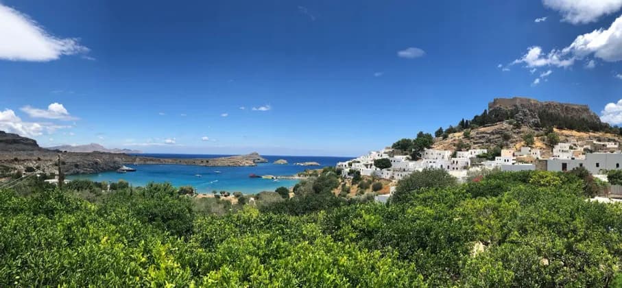 Un villaggio imbiancato a calce e un'acropoli su un pendio che domina una baia turchese, con alberi verdi in primo piano.