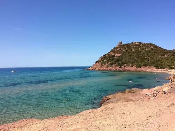 Una torre di pietra su una collina verde si affaccia su una baia calma con acqua turchese, una barca a vela e persone che nuotano da una spiaggia sabbiosa.