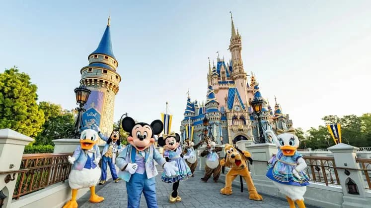 Mickey Mouse y amigos con atuendo formal posan juntos en un puente con un gran castillo de cuento de hadas de fondo.