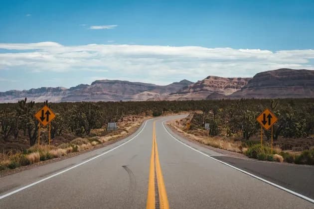 Una strada a due corsie serpeggiante che attraversa un paesaggio desertico con alberi di Joshua e montagne rocciose sotto un cielo parzialmente nuvoloso.
