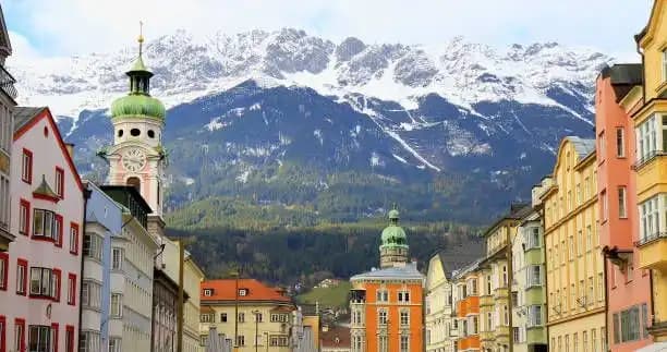 Eine Stadtstraße mit farbenfrohen Gebäuden und einem Uhrturm, vor der Kulisse großer, schneebedeckter Berge.